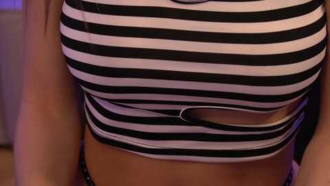 Media: Video of a woman's midsection, wearing a tight, black-and-white striped crop top with a cut-out design. Her skin is tan, and the background is blurred, featuring soft lighting and a hint of purple.