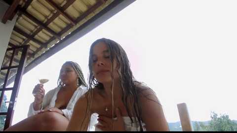 Media: Video of two women with wet hair, one holding a straw, on a wooden deck with a rustic roof, under a bright sky, with a green background.