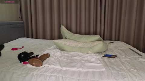 Media: A video of a bed with white sheets, featuring a pile of light green pillows, two vibrators, and a remote control on the bed. Brown curtains and a grey headboard are in the background.