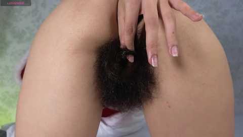 Media: Video of a light-skinned woman with a trimmed pubic area, fingers spreading her labia, showing her vaginal opening. She has long, manicured nails. Background is blurred.