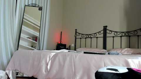 Media: Video of a cozy bedroom with two twin beds covered in pink satin sheets, a black wrought-iron headboard, a tall mirror, and white curtains.