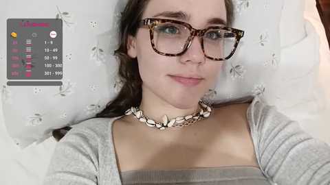 Media: Video of a young woman with light skin, long brown hair, wearing glasses, a gray top, and a silver necklace, against a floral-patterned background.