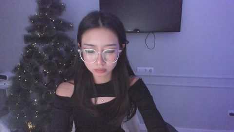 Media: Video of an Asian woman with long black hair, wearing glasses and a black off-shoulder dress, seated indoors beside a lit Christmas tree, looking slightly downwards.