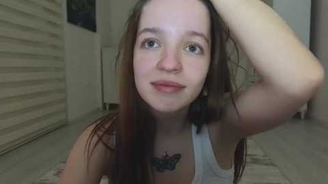 Media: Video of a young Caucasian woman with fair skin, long brown hair, and blue eyes, wearing a white tank top, standing indoors with a closed white door and light-colored tiled floor.