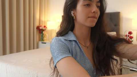 Media: Video of a young South Asian woman with long black hair, wearing a light blue polka-dot blouse, smiling while brushing her hair in a modern, well-lit bedroom.