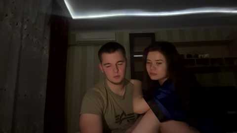 Media: Video of a young couple, man with short hair in a beige t-shirt, woman with long dark hair in a blue top, sitting on a bed in a dimly lit room with patterned wallpaper and a ceiling light.