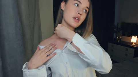 Media: Video of a young Caucasian woman with light skin and shoulder-length brown hair, in a dimly lit bedroom, adjusting her white button-up shirt.