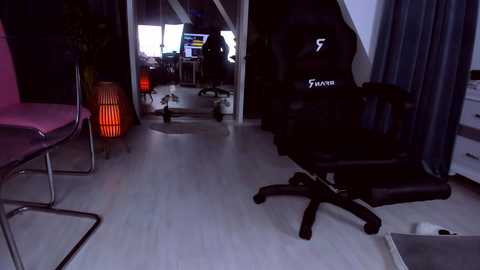 Media: Video of a dimly lit gaming room with a black gaming chair, desk, and monitors; a red lamp adds warmth.