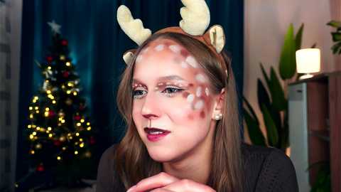 Media: Video of a young woman with fair skin, wearing reindeer antlers, white face paint, and red lipstick, smiling, in a cozy room with a Christmas tree and lit candles.