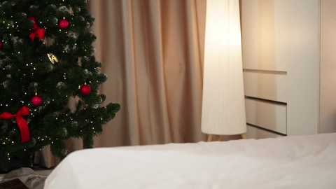 Media: Video of a cozy bedroom scene featuring a lit Christmas tree with red baubles and a gold star, beside a white lamp, and a white bed, with beige curtains and a white dresser in the background.