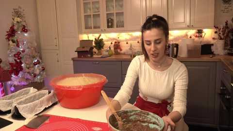 Media: Video of a woman in a white long-sleeve top and red apron mixing brown cake batter in a bowl on a kitchen island. Background features a Christmas tree, cabinets, and countertops.