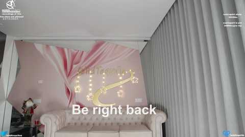 Media: Video of a modern, light-filled room with a pink and white floral mural on the wall, a large gold \"M\" sign, white couch, and vertical grey curtains. Text overlay reads \"Be right back.\