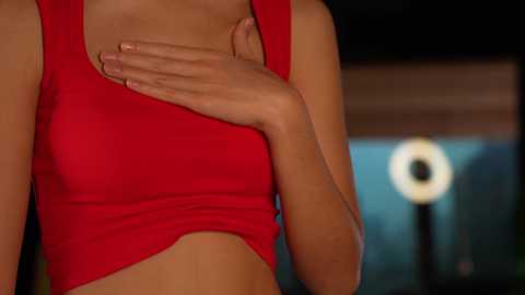 Media: Video of a woman in a red tank top, hands on chest, blurred background with a bright light source.