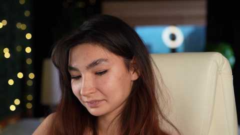 Media: Video of a young Asian woman with long, dark hair, light skin, and closed eyes, sitting in a modern, cream-colored chair. The background features a dark, blurred room with fairy lights and a large ring light.