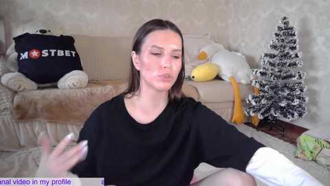 Media: Video of a young woman with light skin, straight brown hair, wearing a black T-shirt, applying makeup, sitting on a beige sofa with stuffed toys and a Christmas tree in a cozy living room.