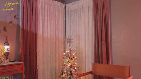 Media: A video of a cozy room with a decorated Christmas tree in a corner, surrounded by sheer white and maroon curtains, and a wooden chair.