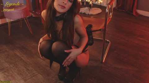 Media: Video of a woman in black lingerie and thigh-high stockings, squatting seductively in a dimly lit room with red lighting, wooden floor, and modern furniture.