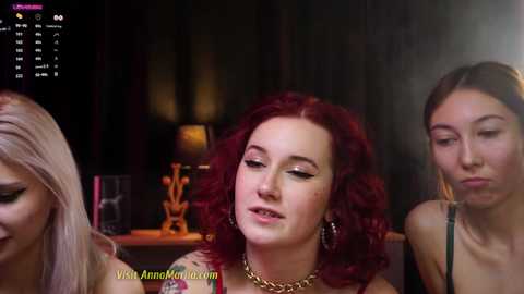 Media: Video of three women with different hair colors and makeup styles sitting closely, smiling, in a dimly-lit room with a calendar and a lamp in the background.