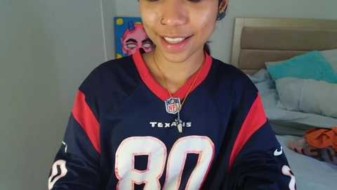 Media: Video of a smiling woman wearing a Dallas Cowboys jersey, standing in a bedroom with a bed, blue wall, and colorful artwork.