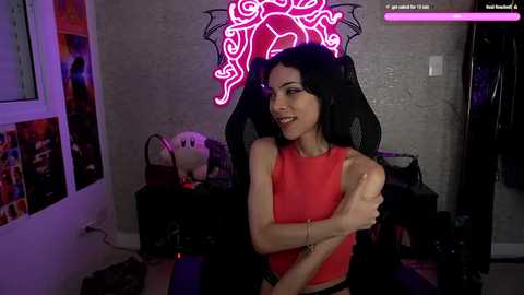 Media: Video of a smiling, slender woman with long black hair, wearing a sleeveless red top, sitting in a black gaming chair. Neon pink light illuminates the background.