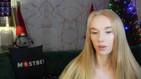 Media: Video of a blonde woman with long hair, light skin, and full lips, sitting on a green couch. She's wearing a black top. Christmas decorations in the background.