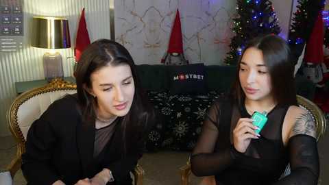 Media: Video of two young women in a cozy living room; one with long dark hair, black sweater, and tattoos, the other with straight dark hair, black top, holding a green card.