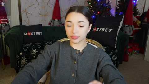 Media: Video of a young girl with dark hair, wearing a gray sweater, sitting in a living room with a green couch and a decorated Christmas tree.