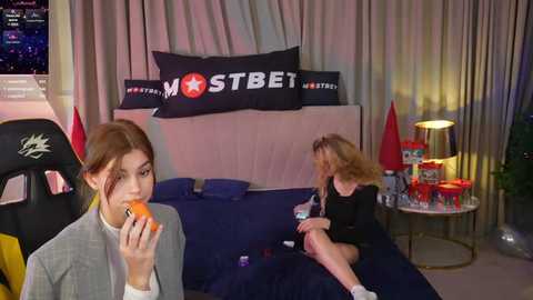 Media: Video of two young women, one with an orange in her mouth, sitting on a bed in a dimly lit room with a \"MST3K\" banner, gaming equipment, and drinks.