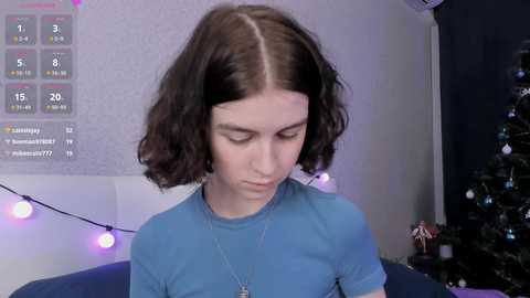 Video of a young woman with shoulder-length brown hair, wearing a blue t-shirt, standing indoors near a decorated Christmas tree.