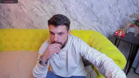 Media: A video of a bearded man in a white jacket, sitting on a bright yellow sofa, against a textured gray wall, with a black side table and festive decorations in the background.