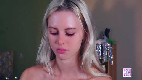 Media: Video of a young, fair-skinned, blonde woman with long hair, wearing a top, looking down thoughtfully. Background features a blurred green wall and a glittery ornament.