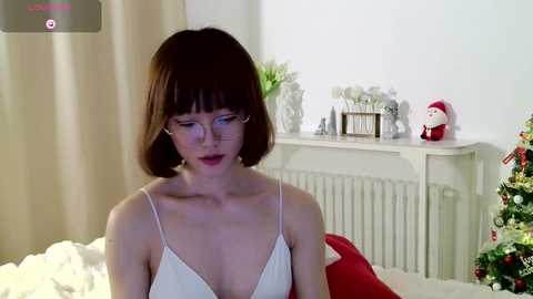 Media: Video of a fair-skinned woman with bob-cut hair in a white camisole, sitting in a cozy bedroom with a Christmas tree and festive decorations.