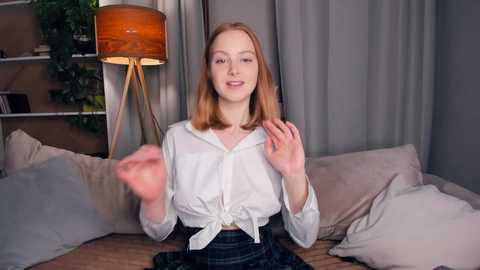 Media: A video of a young woman with fair skin and shoulder-length red hair, smiling, wearing a white blouse tied at the waist and a black skirt, seated on a beige sofa. Background features a lamp, greenery, and light grey curtains.