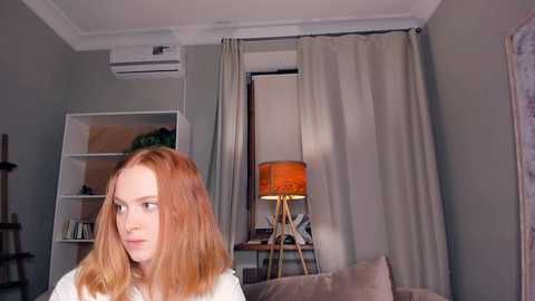 Media: Video of a red-haired woman with fair skin in a modern living room, featuring beige walls, a window with beige curtains, a wooden floor lamp, and a white bookshelf.