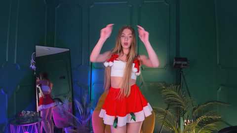 Media: Video of a young, slender, Caucasian woman with long blonde hair in a red and white Santa-themed crop top and skirt, dancing energetically in a dimly lit, green-themed room with studio equipment and greenery in the background.