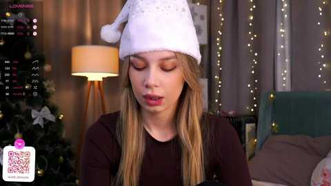 Media: Video of a young blonde woman in a white Santa hat, wearing a dark shirt, indoors, with a Christmas tree and string lights in the background.