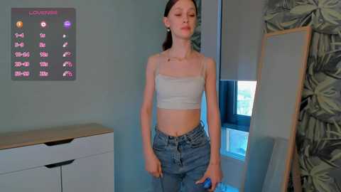 Media: Video of a young woman with fair skin, brown hair, wearing a white crop top and high-waisted jeans, standing indoors in a room with a clock on the wall and a leaf-patterned door.