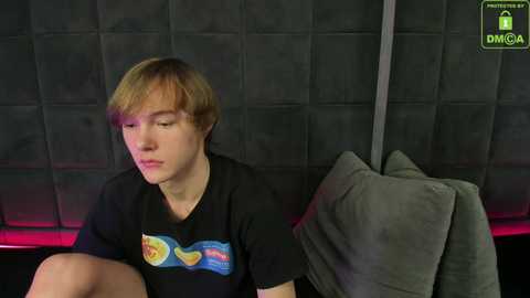 Media: Video of a young, fair-skinned, light-haired boy with short hair, wearing a black T-shirt with a colorful graphic, sitting on a gray cushion against a dark, padded wall in a dimly lit room.