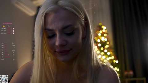 Media: A dimly lit video of a blonde woman with fair skin, wearing a sleeveless top, standing in front of a Christmas tree with warm, glowing lights in a softly lit room.