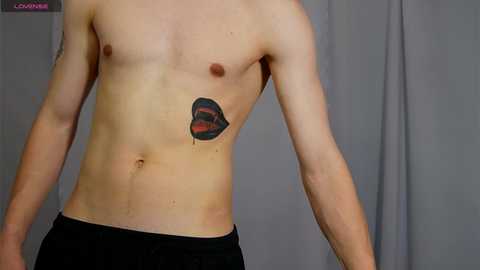 Media: Video of a shirtless, fair-skinned, slender male with a small, red, black, and green tattoo of a lips on his left chest. He wears black pants, standing against a gray curtain backdrop.