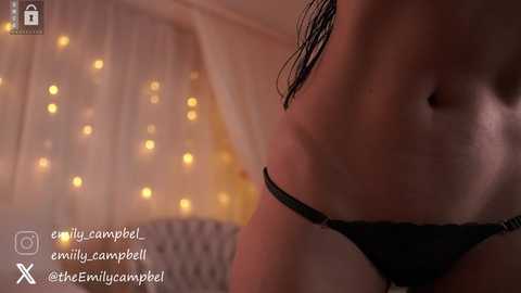 Video of a woman's torso in black underwear, with a dimly lit background featuring string lights and sheer curtains.