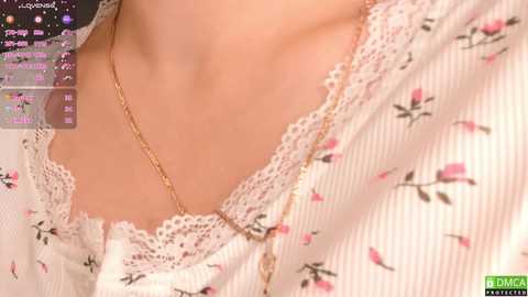 Media: Video of a woman's neck and chest, wearing a white lace-trimmed blouse with pink floral patterns. She has a delicate gold necklace. Background shows a computer screen displaying a calendar and various icons.