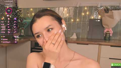 Media: A video of a young woman with light skin and brown hair, wearing white headphones and a black wristwatch, covering her mouth with her hand, in a modern kitchen with a tiled backsplash and fairy lights.