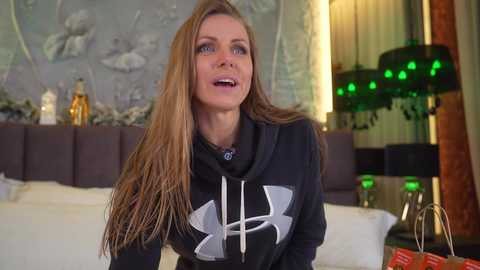 Media: Video of a fit, blonde woman in a black Under Armour hoodie, with long straight hair, talking animatedly in a modern, cozy living room with a floral wall, green LED lights, and a black lamp.