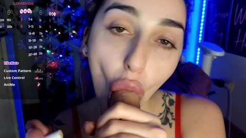 Media: Video of a young woman with fair skin and dark hair, performing oral sex on a large erect penis. Background features Christmas decorations and blue LED lighting, suggesting a festive indoor setting.