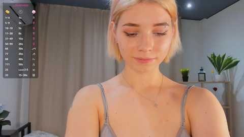 Media: Video of a fair-skinned woman with short blonde hair in a grey tank top, standing indoors. Background includes beige curtains, a plant, and a calendar.