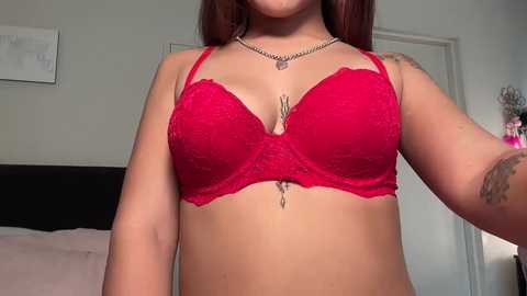 Media: Video of a woman with light brown skin, wearing a red lace bra, revealing medium-sized breasts, and a tattoo on her right arm. Background shows a white door, a bed, and a pink vase.