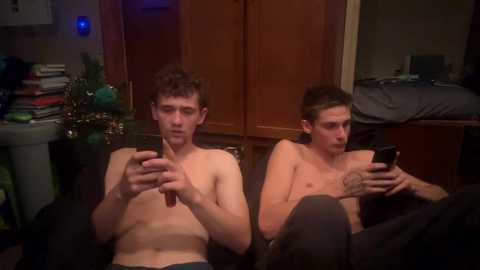 Media: Video of two shirtless, slender young men with short hair, sitting on a couch, holding phones, in a dimly lit room with wooden cabinets and cluttered bookshelf.