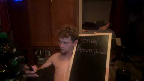 Media: Video of a shirtless, curly-haired young man with light skin, writing on a chalkboard. Background features a wooden kitchen, partially visible, with a Christmas tree adorned with ornaments to the left.