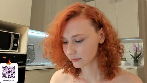 Media: Video of a pale-skinned, young woman with shoulder-length, curly, fiery red hair, standing in a modern kitchen with white cabinets, a microwave, and a potted plant.